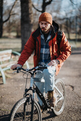 Focused young adult wearing a beanie and jacket bicycles leisurely on a sunny day in the park, showcasing an active lifestyle.