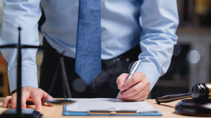 A lawyer in a blue shirt and black pants is writing and signing on a piece of paper document with a...