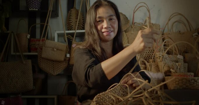 A smiling Asian female artisan who is a rural villager weaving folk women's baskets and bags made from leftover paper band to sell as extra income in the community shop.