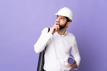 Young architect man with helmet and holding blueprints over isolated purple background having...