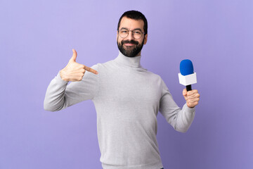 Adult reporter man with beard holding a microphone over isolated purple background proud and...