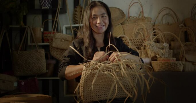 A smiling Asian female artisan who is a rural villager weaving folk women's baskets and bags made from leftover paper band to sell as extra income in the community shop.