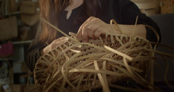 An Asian female artisan who is a rural villager weaving folk women's baskets and bags made from leftover paper band to sell as extra income in the community shop.