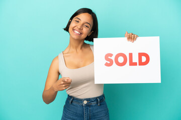 Young mixed race woman isolated on blue background holding a placard with text SOLD and pointing to...