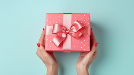 Female hands with red manicure hold a pink gift box with a pink ribbon, on a turquoise background,...