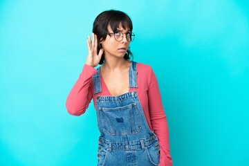 Young woman with overalls isolated background listening to something by putting hand on the ear