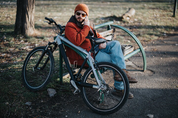 A relaxed young man in casual attire sits with his bicycle in a park, enjoying a serene moment in...