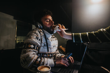 A dedicated man multitasking in a dimly lit office space, eating a snack with one hand while using a smart phone and working on a laptop. - obrazy, fototapety, plakaty
