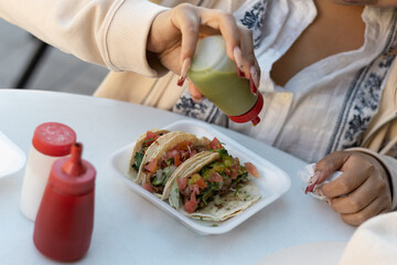 Tourist woman ready to eat meat tacos with vegetables and is seasoning them with spicy green sauce in an outdoor Mexican food restaurant; shopping customer, buying consumables