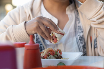 Photograph of the hands of a female tourist holding a meat and vegetable taco that she is enjoying at a local Mexican restaurant, inside an outdoor grocery market