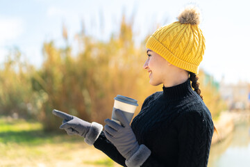 Young moroccan girl wearing winter muffs while holding a coffee at outdoors pointing to the side to...