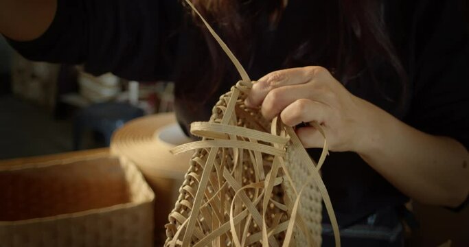 An Asian female artisan who is a rural villager weaving folk women's baskets and bags made from leftover paper band to sell as extra income in the community shop.