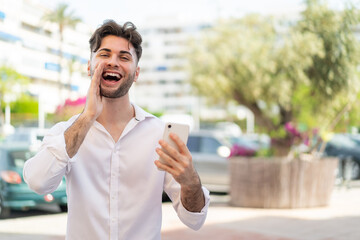 Young handsome man using mobile phone at outdoors shouting with mouth wide open
