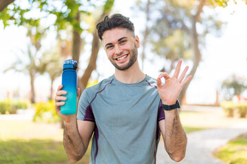 Young handsome sport man with a bottle of water at outdoors showing ok sign with fingers