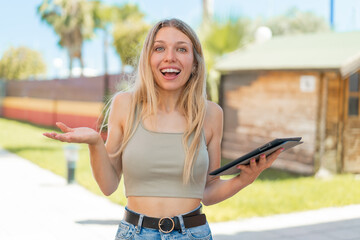 Young blonde woman holding a tablet at outdoors with shocked facial expression