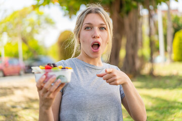 Young blonde woman holding a bowl of fruit at outdoors surprised and pointing front