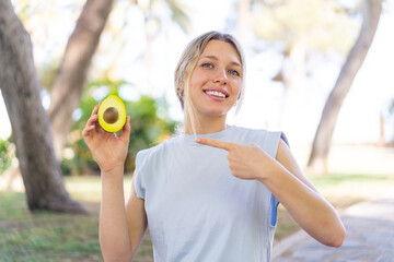 Young blonde woman holding an avocado at outdoors and pointing it
