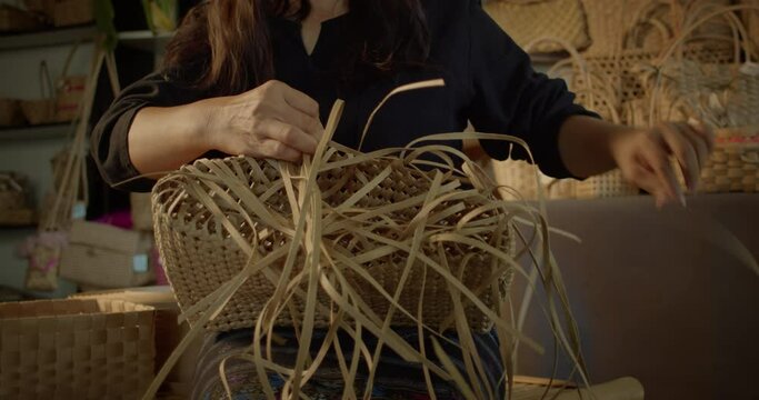 An Asian female artisan who is a rural villager weaving folk women's baskets and bags made from leftover paper band to sell as extra income in the community shop.