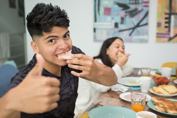 Young adult eating breakfast looking at camera at home happy with thumb up