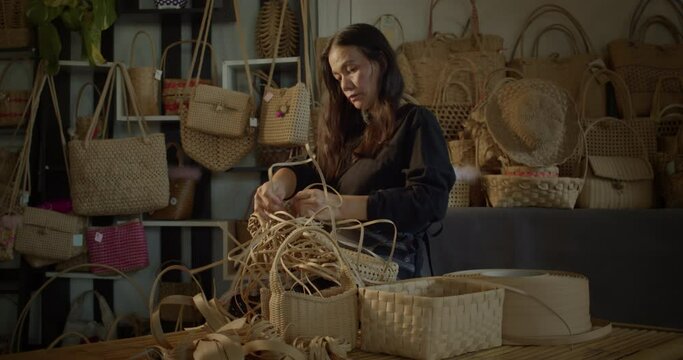 An Asian female artisan who is a rural villager weaving folk women's baskets and bags made from leftover paper band to sell as extra income in the community shop.