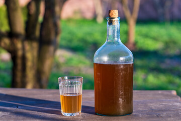 Big bottle with a drink made from fermented birch sap on the wooden table on a warm spring day, closeup. Traditional Ukrainian cold barley drink kvass in a glass jar and glass in yard - obrazy, fototapety, plakaty