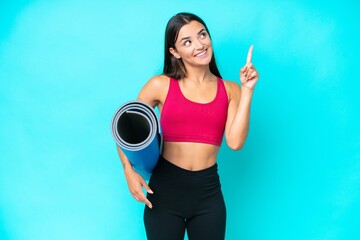 Young sport caucasian woman going to yoga classes while holding a mat isolated on blue background pointing up a great idea