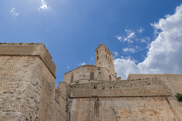 An upward view of the Eivissa Almudaina Castle in Ibiza, showcasing its historic stone walls and robust clock tower against a backdrop of scattered clouds in a blue sky