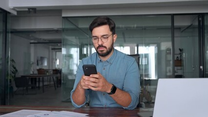 Busy happy young latin business man holding smartphone standing in office. Professional hispanic businessman entrepreneur or manager using financial banking apps on cell phone technology at work. - Powered by Adobe
