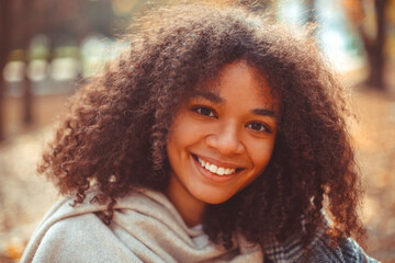 Cute autumn close up portrait of young smiling happy african american woman with curly hair enjoying walk in park in fall season