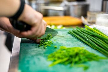 Close-up of a chef’s hands slicing fresh green onion on a green cutting board in a professional kitchen, with sliced onion and utensils in the background