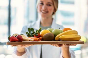 Beautiful smiling nutritionist holding wooden table with fresh fruits and vegetables in a medical consultation