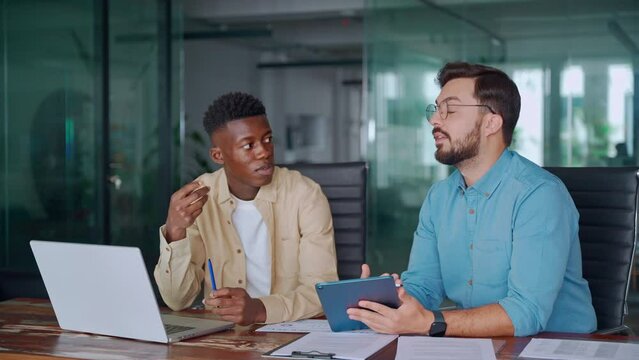 Two busy diverse male employees using laptop and tablet talking in office. Professional young business men workers team having conversation working on project sharing ideas sitting at work desk.