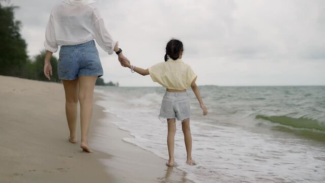 A woman holds a child's hand while walking on the beach. The child wore a yellow shirt and blue jeans. The scene is calm and relaxing. with the ocean in the background