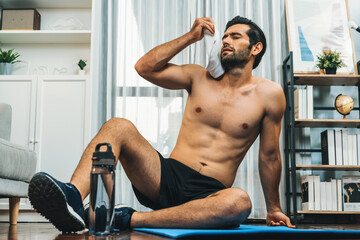 Athletic and sporty man sitting, resting on fitness mat after finishing home body workout exercise...