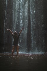 Vertical shot of a woman standing with hands up in front of a big waterfall