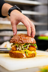 Hand placing the top bun on a gourmet burger with fried chicken, cheese, and fresh vegetables, in a professional kitchen setting