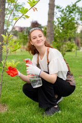 Front view of girl spraying trees, holding bottle, looking at camera, smiling. Concept of taking care of plants.