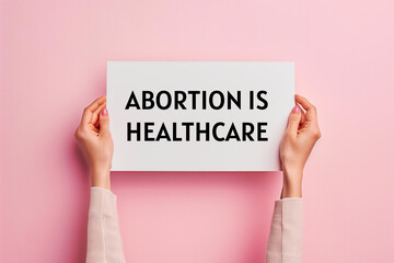 photo womans hands holding up a banner with text abortion is healthcare on a pastel pink background