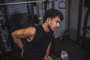 Naklejka premium A young asian man suffers from lower back pain while working out at the gym. Poor posture or overuse leading to chronic injury.