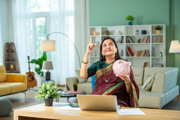 Finance concept - Indian retired old lady holding piggy bank, money, laptop and 3d home model at home