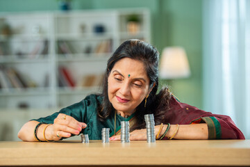 Indian asian senior woman making towers of money coins on table, showing growth or returns on...