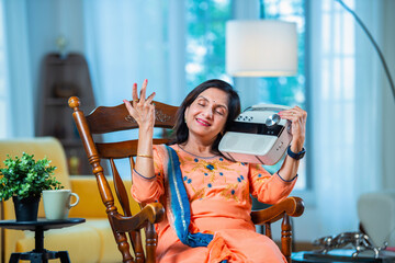 Indian senior adult woman having fun listening to radio at home while sitting on rocking chair.