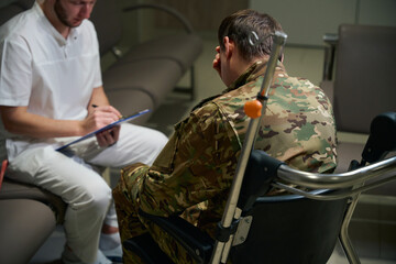Focused doctor writing down complaints of serviceman with disability - obrazy, fototapety, plakaty