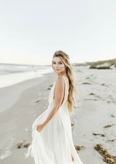 Elegant woman in white dress strolling on the beach at sunset, serene coastal beauty