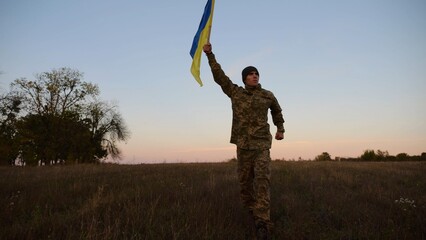 Soldier of ukrainian army running with raised blue-yellow banner on field at dusk. Young male...