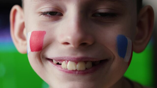 a handsome 10-year-old boy with a French flag painted on his cheek looks at the camera. patriot of France, fan of the national team with the country's symbol painted on his face against the backdrop o