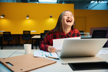 Laughing asian female IT employee working with documents and laptop at desk