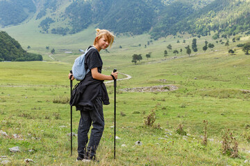 Rear view of hiker female with backpack and trekking pole standing on green lawn on the backdrop of...