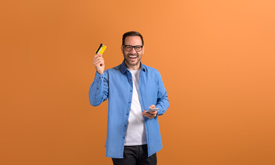 Cheerful young salesman laughing and showing credit card while using smartphone on orange background - obrazy, fototapety, plakaty