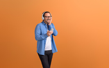 Happy young man with eyes closed enjoying music over headphones and using phone on orange background